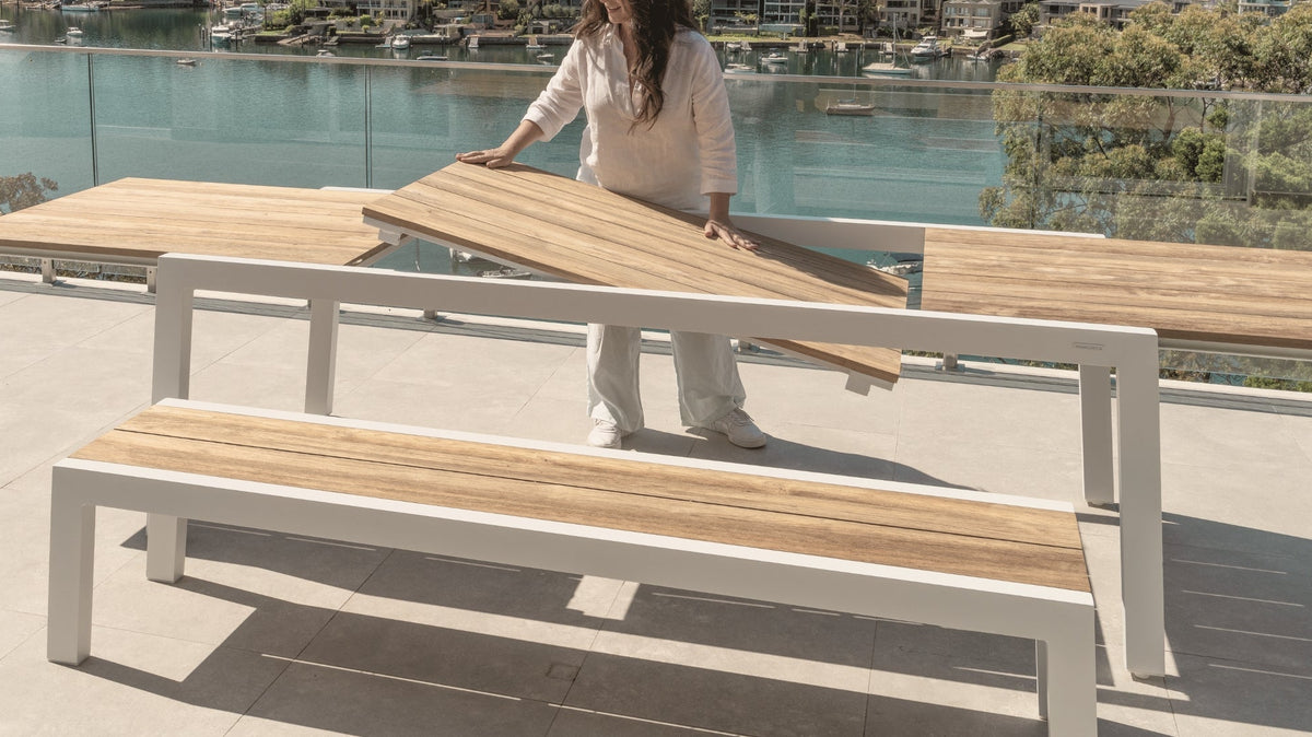 Woman placing the teak extension panel into a white aluminium outdoor dining table, demonstrating the extendable mechanism on a terrace overlooking the water.