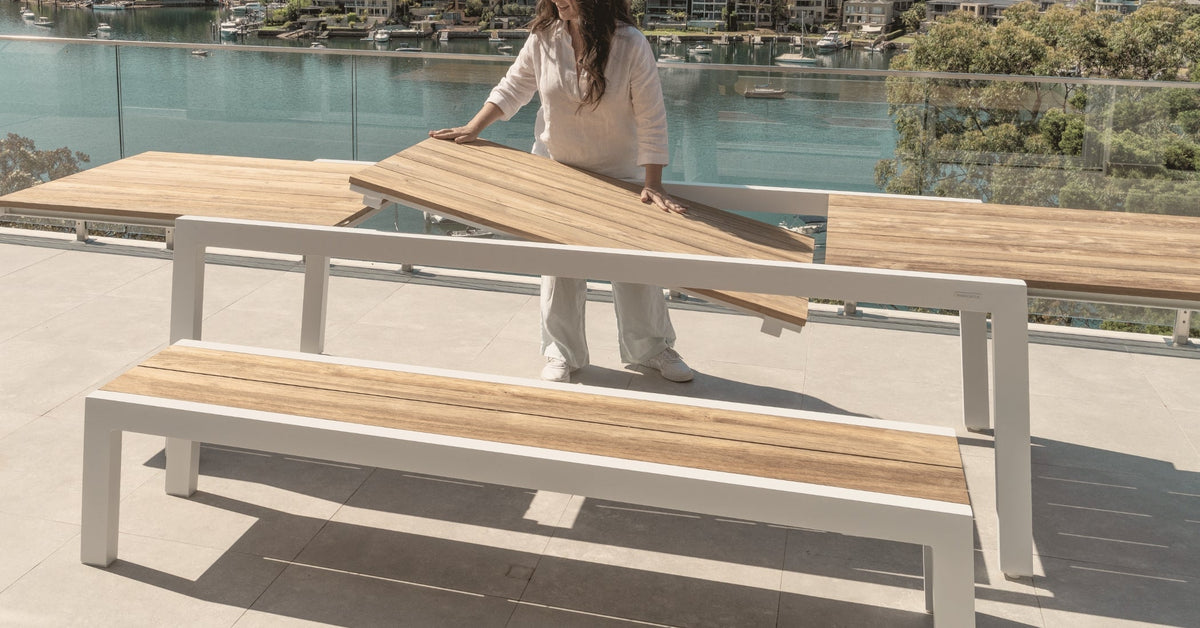Woman placing the teak extension panel into a white aluminium outdoor dining table, demonstrating the extendable mechanism on a terrace overlooking the water.