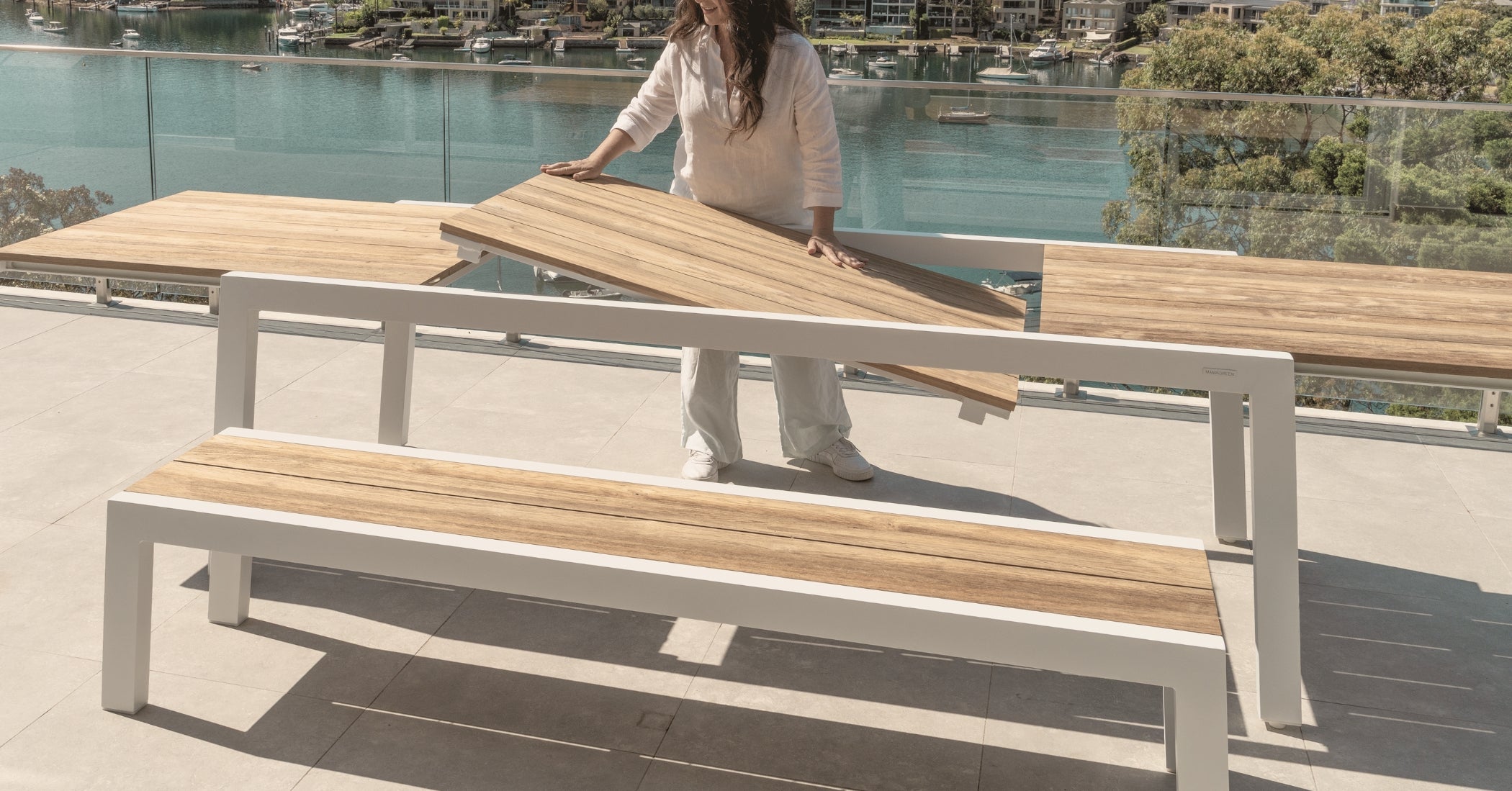 Woman placing the teak extension panel into a white aluminium outdoor dining table, demonstrating the extendable mechanism on a terrace overlooking the water.