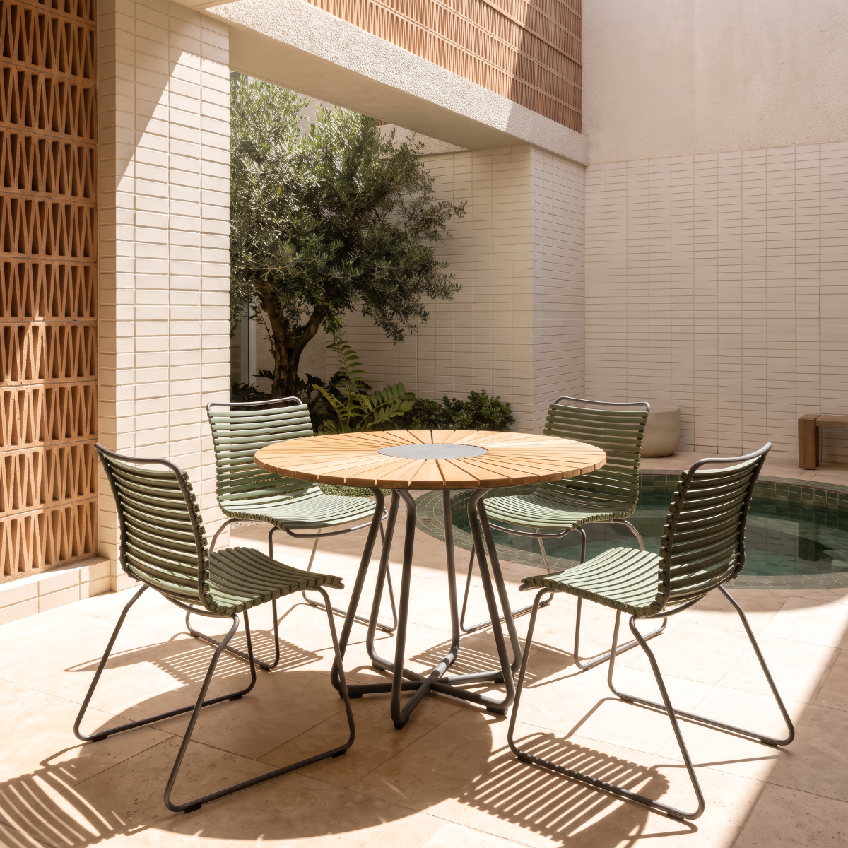 A round wooden outdoor dining table with a slatted metal base, surrounded by four green slatted chairs, set in a modern patio courtyard next to a tiled pool.