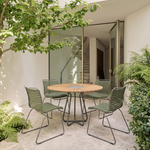 Outdoor patio dining set with a round light wood table and four olive green woven sling chairs, arranged on a stone floor in a modern courtyard with glass doors and greenery.