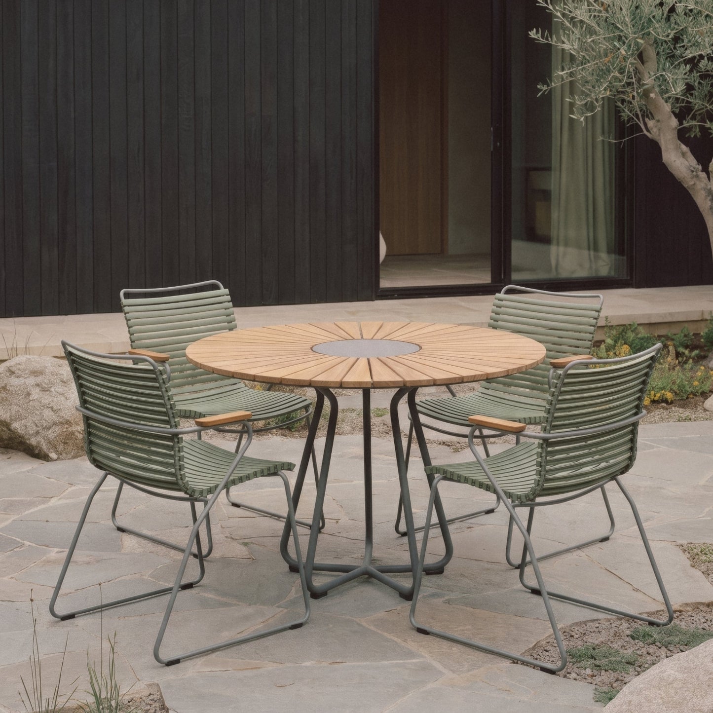 A round outdoor dining table with a light wood top and metal base, surrounded by four green outdoor chairs on a stone patio in front of a modern dark wood building.