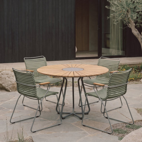 A round outdoor dining table with a light wood top and metal base, surrounded by four green outdoor chairs on a stone patio in front of a modern dark wood building.