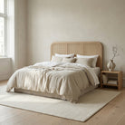 Neatly made bed with beige bedding in a room with a wooden floor and a vase on a nightstand.