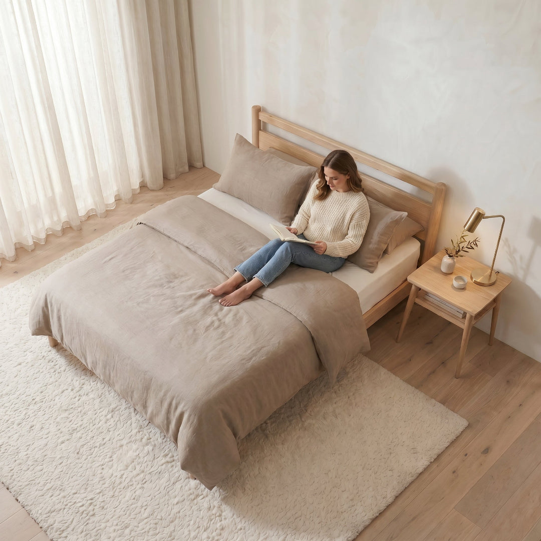 Woman sitting on a bed in a cozy bedroom with beige bedding and wooden furniture.