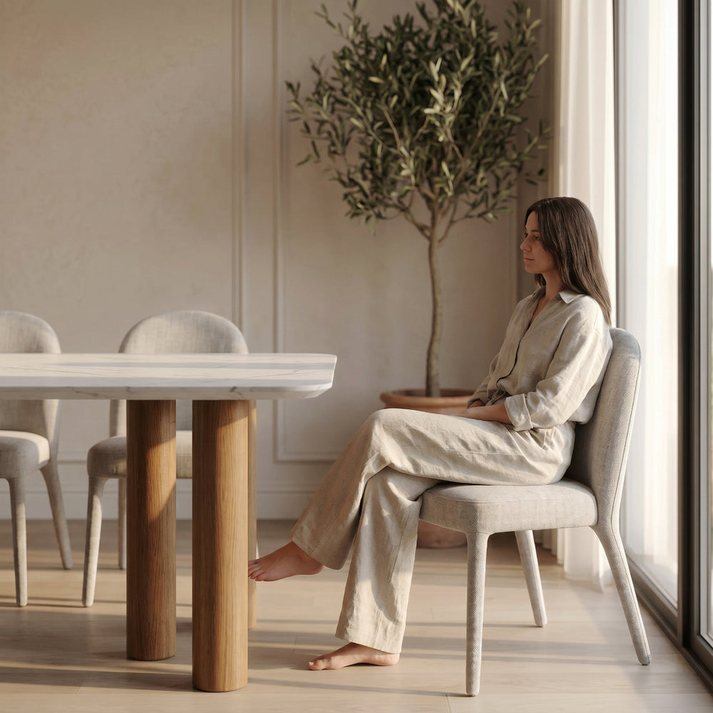 Woman sitting at a dining table in a modern room with a plant and large window.