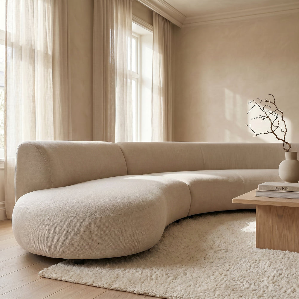 Beige curved sofa in a modern living room with light wood coffee table and large windows.