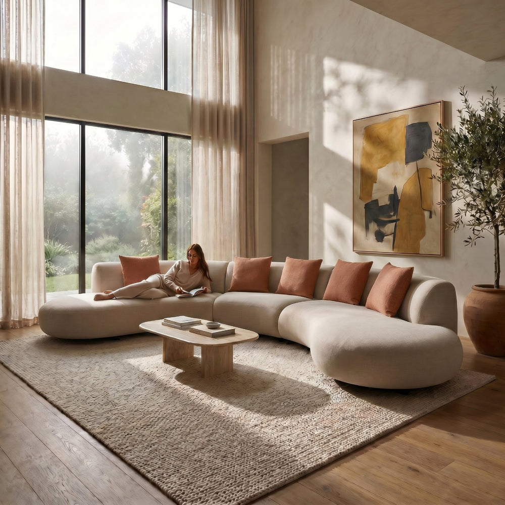 Modern living room with a woman sitting on a sectional sofa, large windows, and decorative elements.