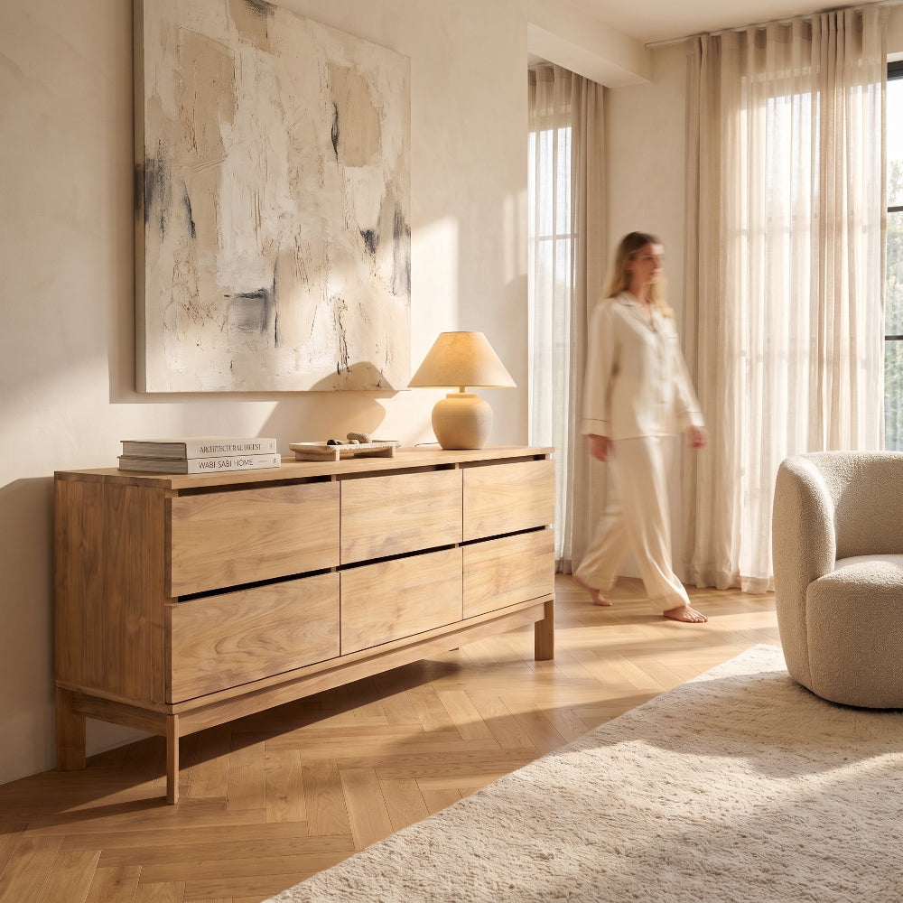 A light oak wood six-drawer dresser in a bright, modern living room. A woman in a matching cream lounge set walks past the dresser, which is styled with a lamp, books, and a decorative bowl.