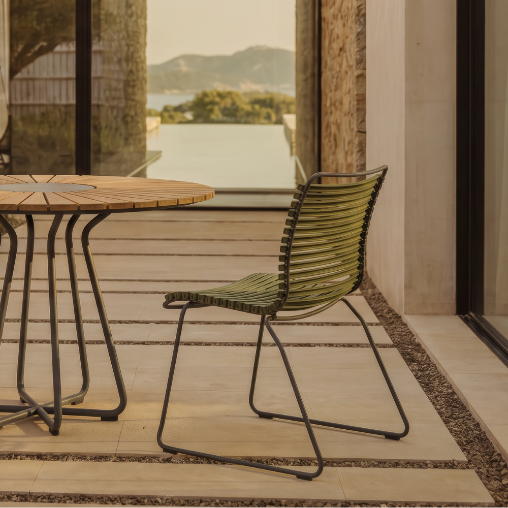 Outdoor patio dining set featuring a round wooden-top table with a black metal frame and two woven green chairs, set on a tiled terrace with a stone wall and large glass windows overlooking a landscape.