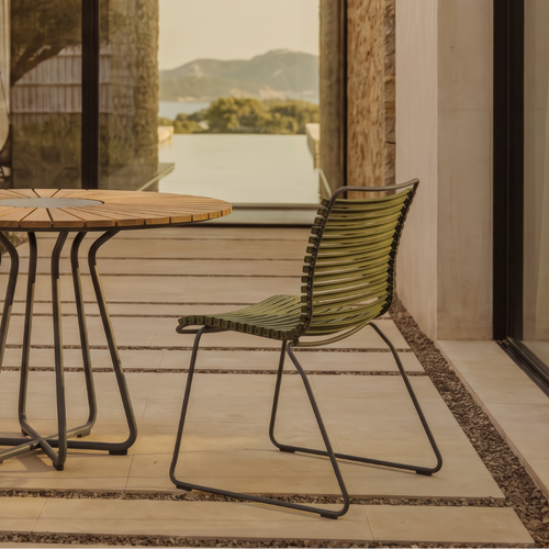 Outdoor patio dining set featuring a round wooden-top table with a black metal frame and two woven green chairs, set on a tiled terrace with a stone wall and large glass windows overlooking a landscape.
