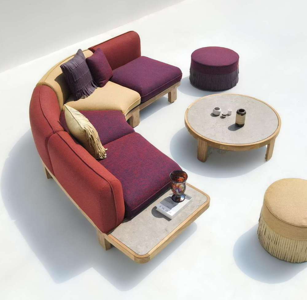 A curved modular outdoor sofa set with red and burgundy cushions, beige fabric sections, and a light wood frame, styled with matching ottomans, a round coffee table, and small decor accessories on a clean white background.