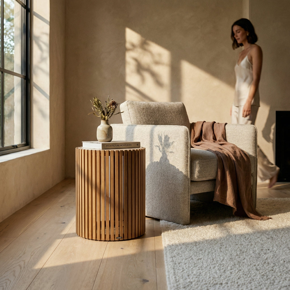 Woman standing in a sunlit room with a beige sofa, wooden side table, and decorative items.